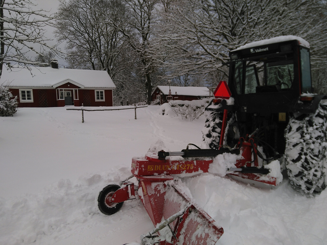 En traktormed skaktblad bakom plogar snö framför ett snötäkt hus. Lövträd i bakgrunden
